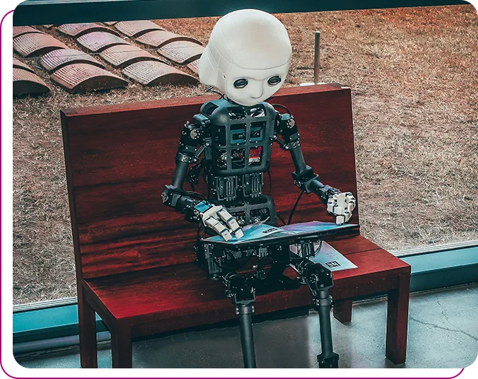 A robot seated at a bench working on a laptop